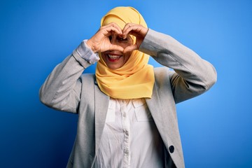 Naklejka premium Middle age brunette business woman wearing muslim traditional hijab over blue background Doing heart shape with hand and fingers smiling looking through sign