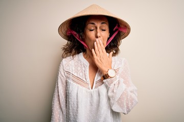 Middle age brunette woman wearing asian traditional conical hat over white background bored yawning tired covering mouth with hand. Restless and sleepiness.