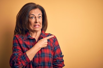 Middle age beautiful woman wearing casual shirt standing over isolated yellow background Pointing aside worried and nervous with forefinger, concerned and surprised expression