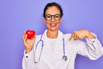 Middle age senior cardiologist doctor woman holding red heart over purple background with surprise...