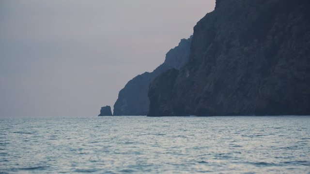 Evening beautiful landscape with rocks, flying seagull and small sea waves. Shot. Natural background of the sunset over the rippled sea surface and the mountains.