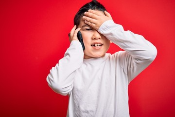 Young little boy kid talking on smartphone mobile over red isolated background stressed with hand on head, shocked with shame and surprise face, angry and frustrated. Fear and upset for mistake.