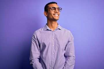 Handsome african american man wearing striped shirt and glasses over purple background looking away to side with smile on face, natural expression. Laughing confident.