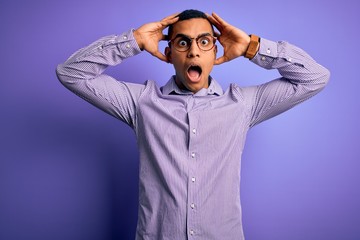 Handsome african american man wearing striped shirt and glasses over purple background Crazy and scared with hands on head, afraid and surprised of shock with open mouth