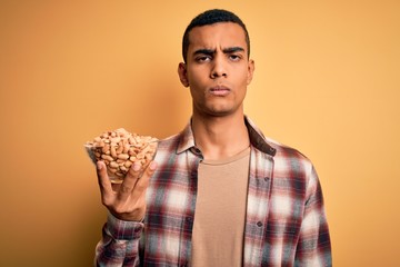 Handsome african american man holding bowl with heathy peanuts over yellow background with a confident expression on smart face thinking serious
