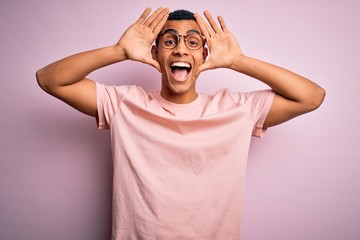 Handsome african american man wearing casual t-shirt and glasses over pink background Smiling cheerful playing peek a boo with hands showing face. Surprised and exited