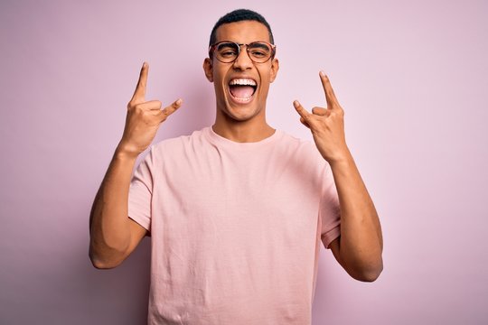 Handsome african american man wearing casual t-shirt and glasses over pink background shouting with crazy expression doing rock symbol with hands up. Music star. Heavy music concept.