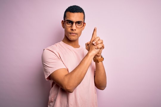 Handsome african american man wearing casual t-shirt and glasses over pink background Holding symbolic gun with hand gesture, playing killing shooting weapons, angry face