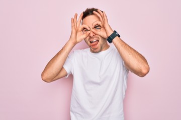 Handsome man with blue eyes wearing casual white t-shirt standing over pink background doing ok gesture like binoculars sticking tongue out, eyes looking through fingers. Crazy expression.