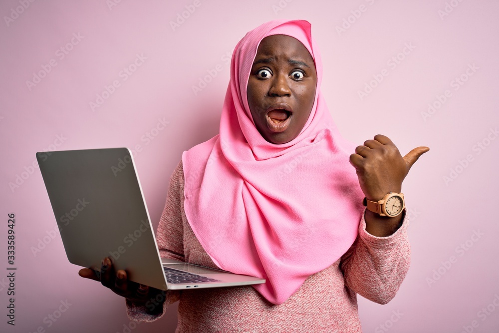 Sticker African american plus size woman wearing muslim hijab using laptop over pink background Surprised pointing with hand finger to the side, open mouth amazed expression.