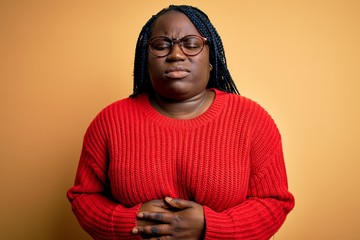 African american plus size woman with braids wearing casual sweater over yellow background with hand on stomach because indigestion, painful illness feeling unwell. Ache concept.