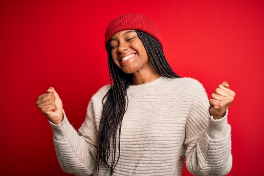 Young african american woman wearing winter sweater and wool hat over red isolated background very happy and excited doing winner gesture with arms raised, smiling and 