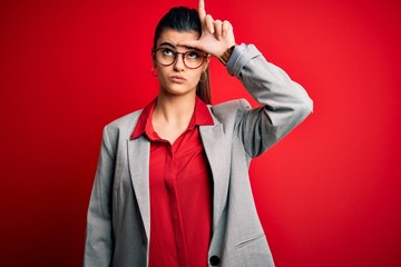 Young beautiful brunette businesswoman wearing jacket and glasses over red background making fun of...