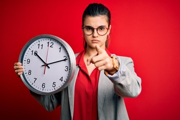 Young beautiful brunette businesswoman doing countdown holding big clock pointing with finger to...