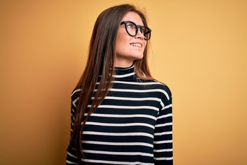 Beautiful woman with blue eyes wearing striped sweater and glasses over yellow background looking away to side with smile on face, natural expression. Laughing confident.