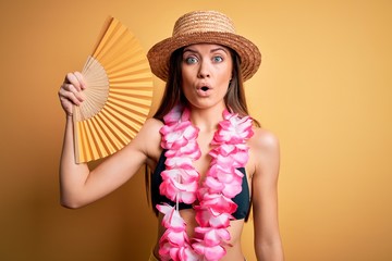 Young beautiful woman with blue eyes on vacation wearing bikini holding hand fan scared in shock with a surprise face, afraid and excited with fear expression