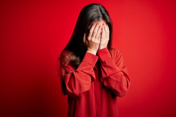 Young beautiful woman with blue eyes wearing casual shirt standing over red background with sad expression covering face with hands while crying. Depression concept.