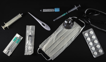 Medical equipment isolated on black background. Surgical protection face mask, syringes, needles, test tube, digital thermometer, pills and stethoscope. Corona Covid-19 protection concept, top view.