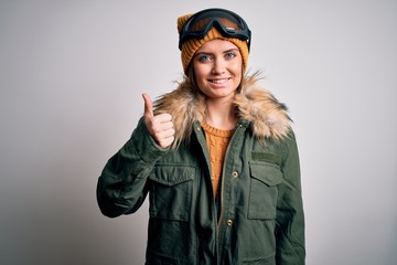 Young beautiful skier woman with blue eyes wearing snow sportswear and ski goggles doing happy thumbs up gesture with hand. Approving expression looking at the camera showing success.