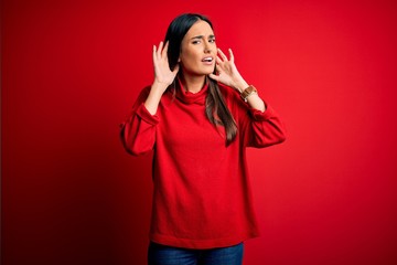 Young beautiful brunette woman wearing casual sweater over isolated red background Trying to hear both hands on ear gesture, curious for gossip. Hearing problem, deaf