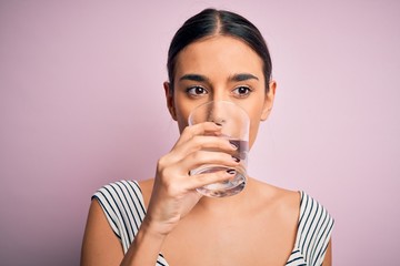 Young beautiful brunette woman drinking glass of healthy water to refreshment standing over isolated pink background