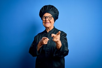 Senior beautiful grey-haired chef woman wearing cooker uniform and hat over blue background...