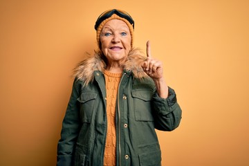 Senior beautiful grey-haired skier woman wearing snow sportswear and ski goggles showing and pointing up with finger number one while smiling confident and happy.