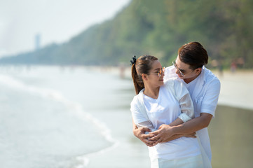 Happy young Asian couple man hugging beautiful woman on tropical beach and looking at each other with smiling face. Healthy family people enjoying relax lifestyle in romantic summer holiday vacation.