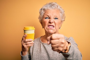 Senior beautiful woman drinking cup of coffee standing over isolated yellow background annoyed and...