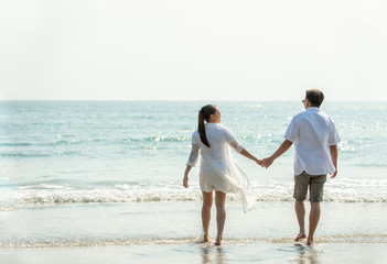 Fototapeta na wymiar Rear view of Happy young Asian couple family man and woman holding hands together and walking on the beach in summertime. Healthy people enjoying relax lifestyle in romantic summer holiday vacation.