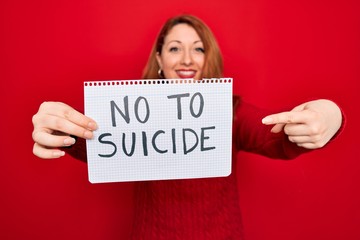 Beautiful redhead woman holding banner showing no to suicide message over red background very happy pointing with hand and finger