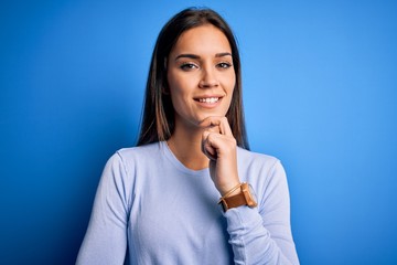 Young beautiful brunette woman wearing casual sweater standing over blue background looking confident at the camera smiling with crossed arms and hand raised on chin. Thinking positive.