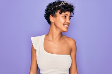 Young beautiful african american afro woman wearing tiara crown over purple background looking away...