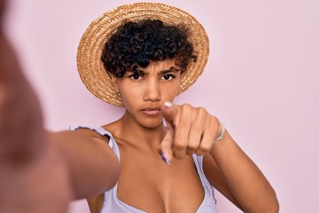 Beautiful african american tourist woman wearing bikini and hat making selfie by camera pointing with finger to the camera and to you, hand sign, positive and confident gesture from the front