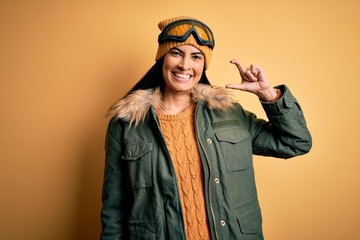 Young beautiful hispanic woman wearing ski glasses and coat for winter weather smiling and confident gesturing with hand doing small size sign with fingers looking and the camera. Measure concept.