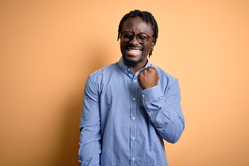 Young handsome african american man wearing shirt and glasses over yellow background celebrating surprised and amazed for success with arms raised and eyes closed. Winner concept.