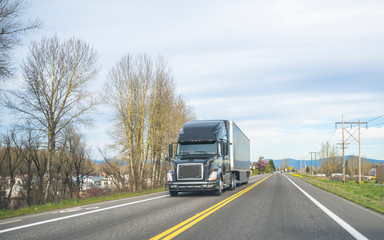 Fototapeta na wymiar Black big rig semi truck transporting cargo in dry van semi trailer running on the two line road