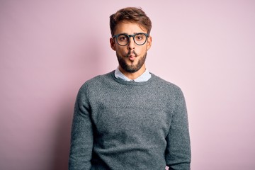 Young handsome man with beard wearing glasses and sweater standing over pink background making fish face with lips, crazy and comical gesture. Funny expression.