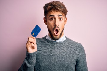 Young customer man with beard holding credit card for payment over pink background scared in shock with a surprise face, afraid and excited with fear expression