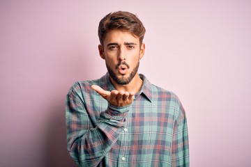 Young handsome man with beard wearing casual shirt standing over pink background looking at the camera blowing a kiss with hand on air being lovely and sexy. Love expression.