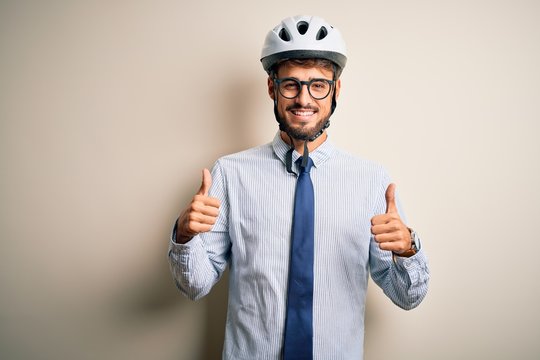 Young businessman wearing glasses and bike helmet standing over isolated white bakground success sign doing positive gesture with hand, thumbs up smiling and happy. Cheerful expression and winner 