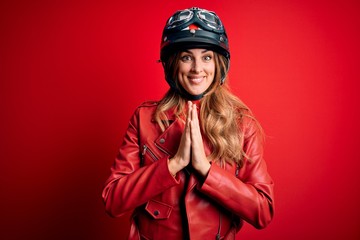 Young beautiful brunette motrocyclist woman wearing moto helmet over red background praying with hands together asking for forgiveness smiling confident.