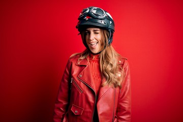 Young beautiful brunette motrocyclist woman wearing moto helmet over red background winking looking at the camera with sexy expression, cheerful and happy face.