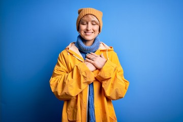 Young blonde woman with short hair wearing rain coat for rainy weather over blue background smiling with hands on chest with closed eyes and grateful gesture on face. Health concept.