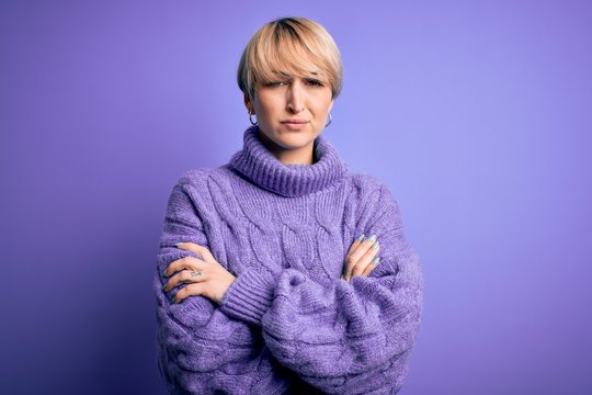 Young blonde woman with short hair wearing winter turtleneck sweater over purple background skeptic and nervous, disapproving expression on face with crossed arms. Negative person.
