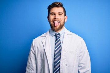 Young blond therapist man with beard and blue eyes wearing coat and tie over background sticking tongue out happy with funny expression. Emotion concept.