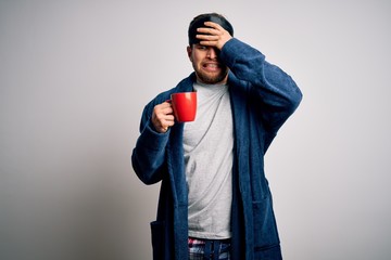 Young blond man with beard and blue eyes wearing pajama drinking cup of coffee stressed with hand on head, shocked with shame and surprise face, angry and frustrated. Fear and upset for mistake.