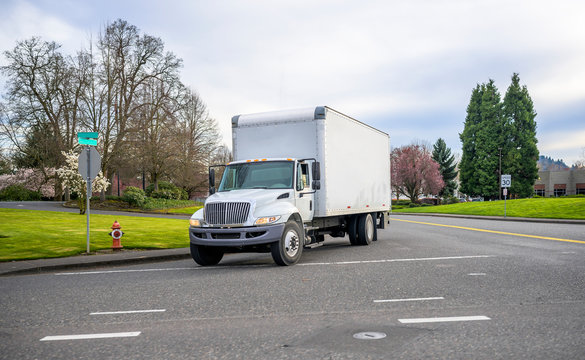 Small compact semi truck with cube box trailer transporting commercial cargo driving on the street of urban city in spring time with blooming trees