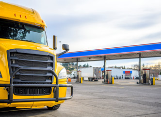 Yellow big rig semi truck with grille guard standing on the truck stop with fuel station on the...