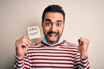 Young handsome man with beard wearing medical mask holding virus alert reminder screaming proud and celebrating victory and success very excited, cheering emotion